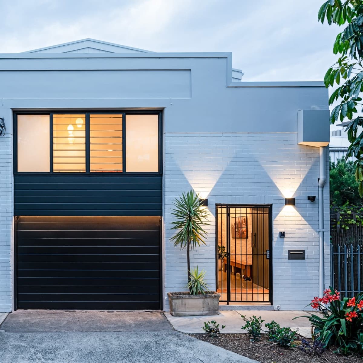 A modern house with a dark garage door, large windows, and tropical plants flanking the entrance.