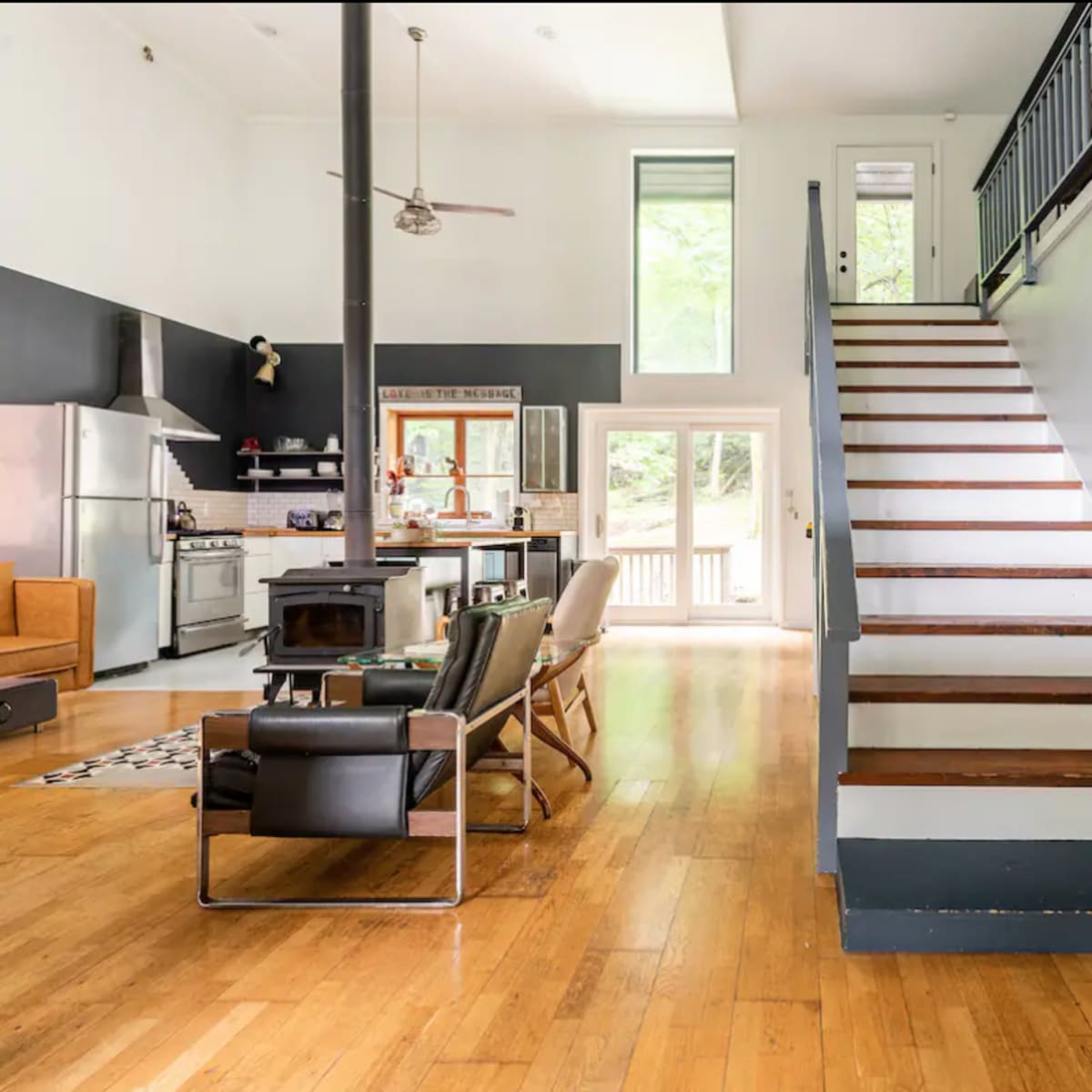 An open-concept living space featuring a kitchen, dining area, and a staircase leading to an upper level, with large windows allowing natural light to enter.