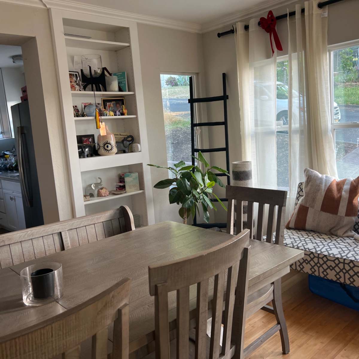 A cozy dining area with a wooden table, chairs, and a window seat adorned with cushions, alongside a small plant and shelves displaying various items.