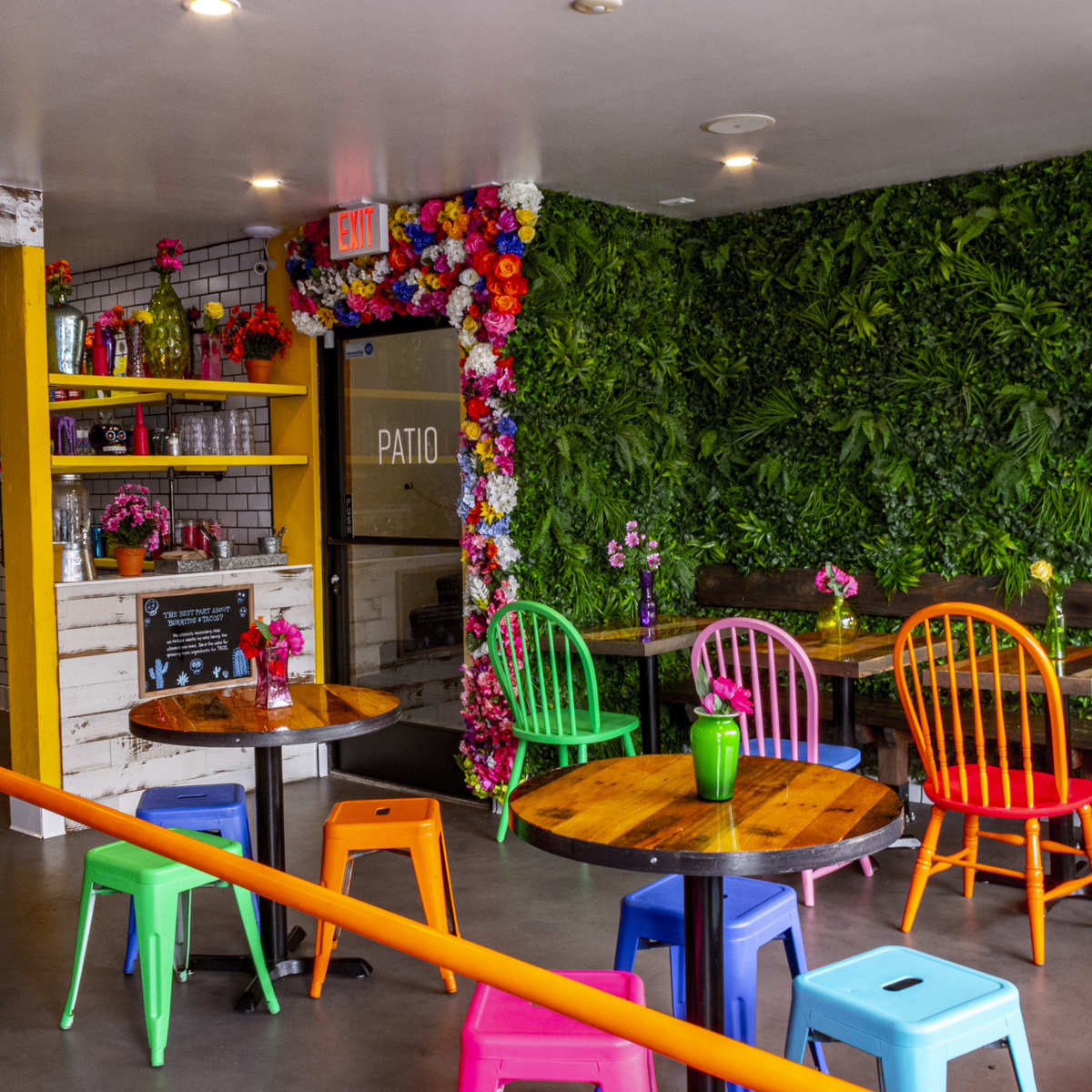 A vibrant café with colorful chairs around wooden tables and a green wall adorned with plants and flowers.