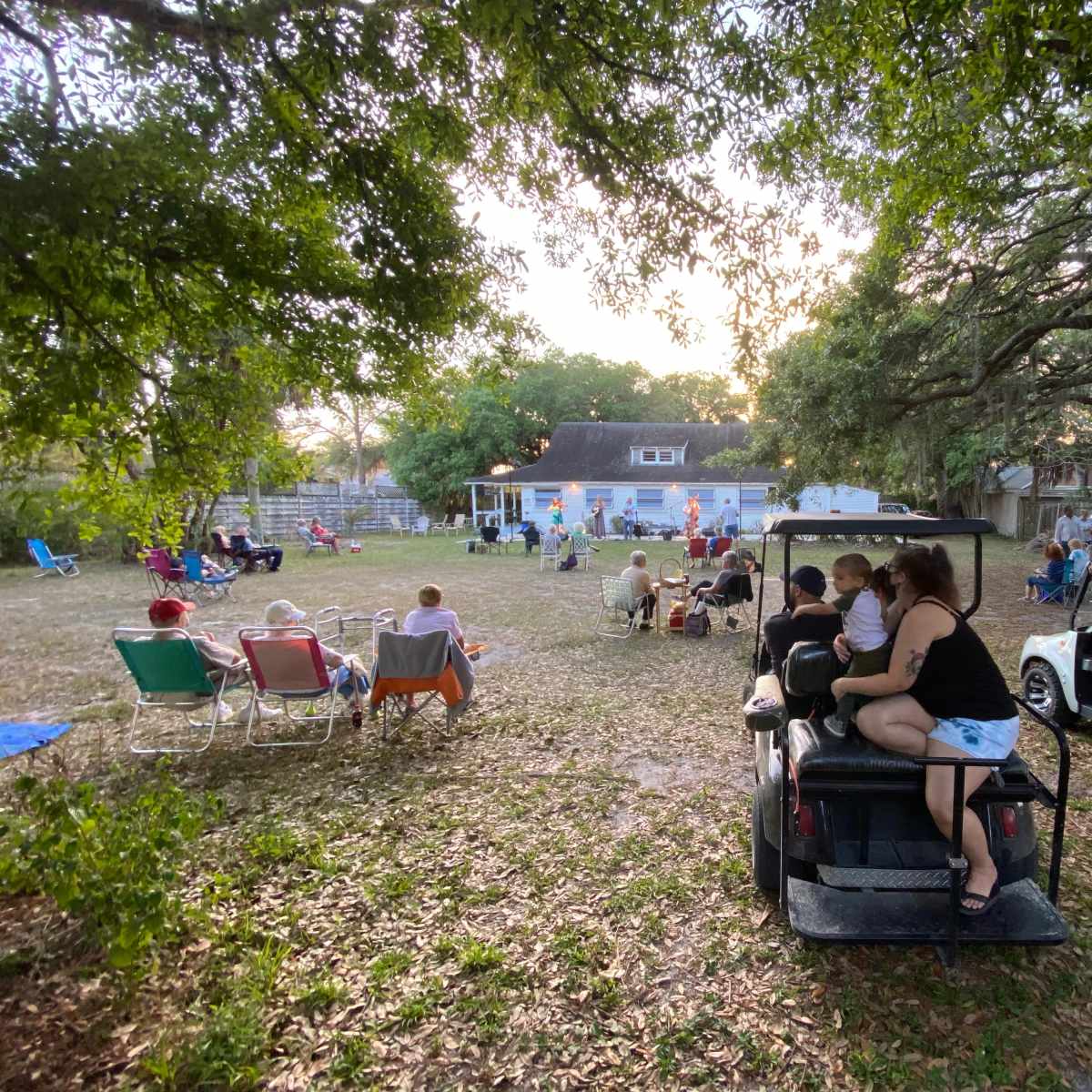 A group of people gather in a grassy area with golf carts parked nearby, while others sit in chairs facing a house in the background.