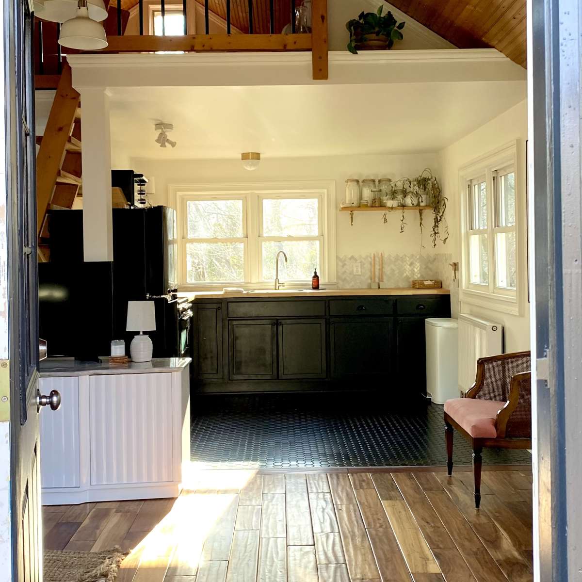 A view of a bright, open kitchen and living area from an entrance, featuring wooden floors, a black kitchen counter, and a staircase leading to a loft.