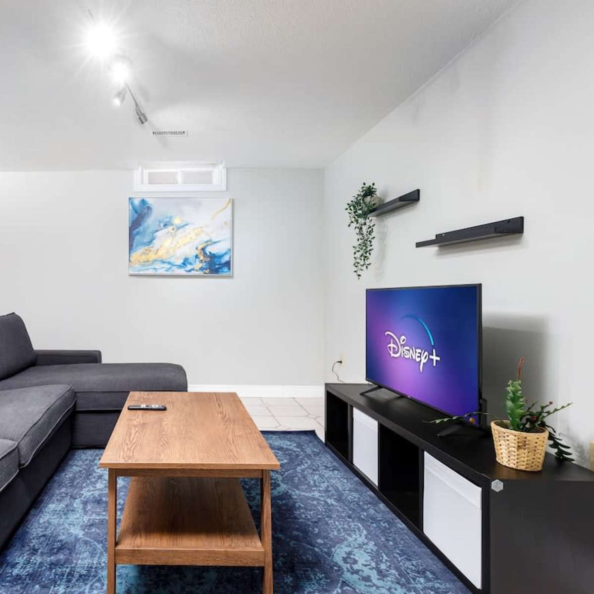 A modern living room features a gray sectional sofa, a wooden coffee table, a television displaying the Disney+ logo, and a painting on the wall.