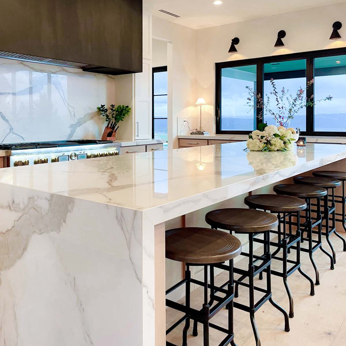 A modern kitchen featuring a marble island with bar stools, a stove, and large windows facing an outdoor view.