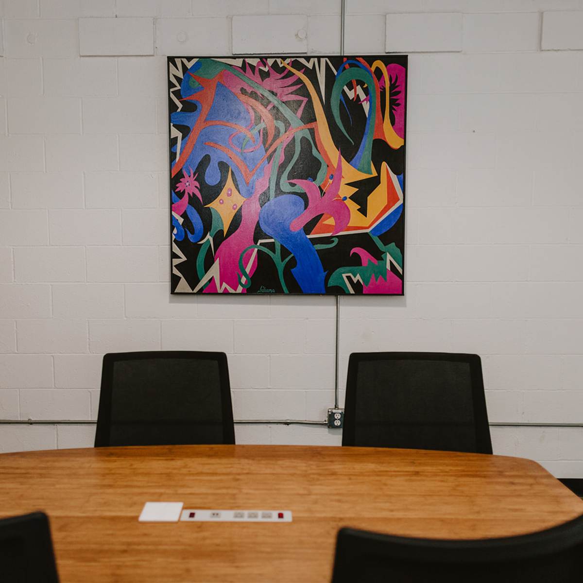 A conference room with a wooden table and three black chairs, featuring a colorful abstract painting on the white wall.