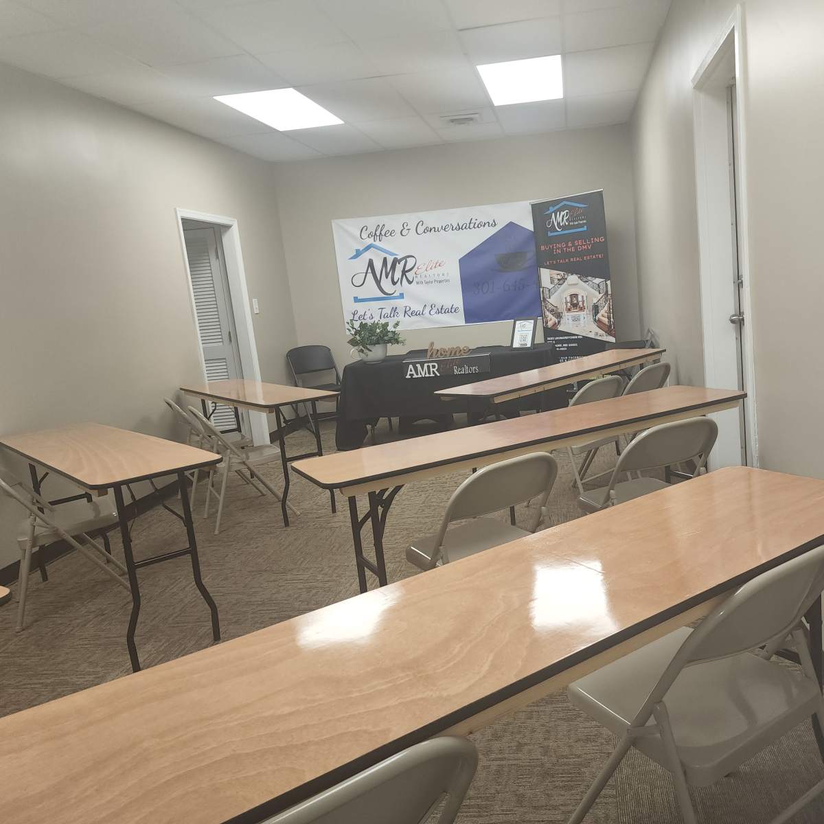 A sparsely furnished meeting room with several wooden tables and chairs arranged in a classroom style, featuring a promotional banner for "Coffee & Conversations" and "AMR" at the front.