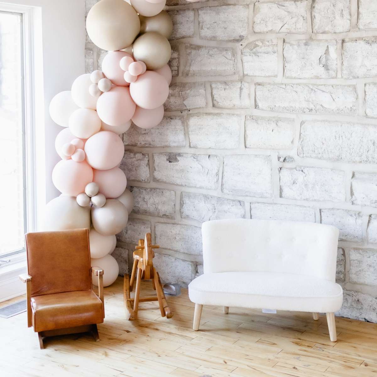 A light-filled room featuring a stone wall decorated with a cluster of balloons in soft colors, alongside a brown armchair and a white sofa on a wooden floor.