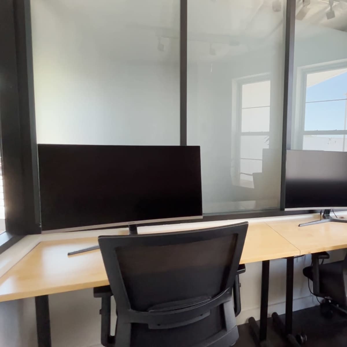A modern office workspace with two black chairs facing two computer monitors on a wooden desk, surrounded by glass walls and a potted plant near a window.