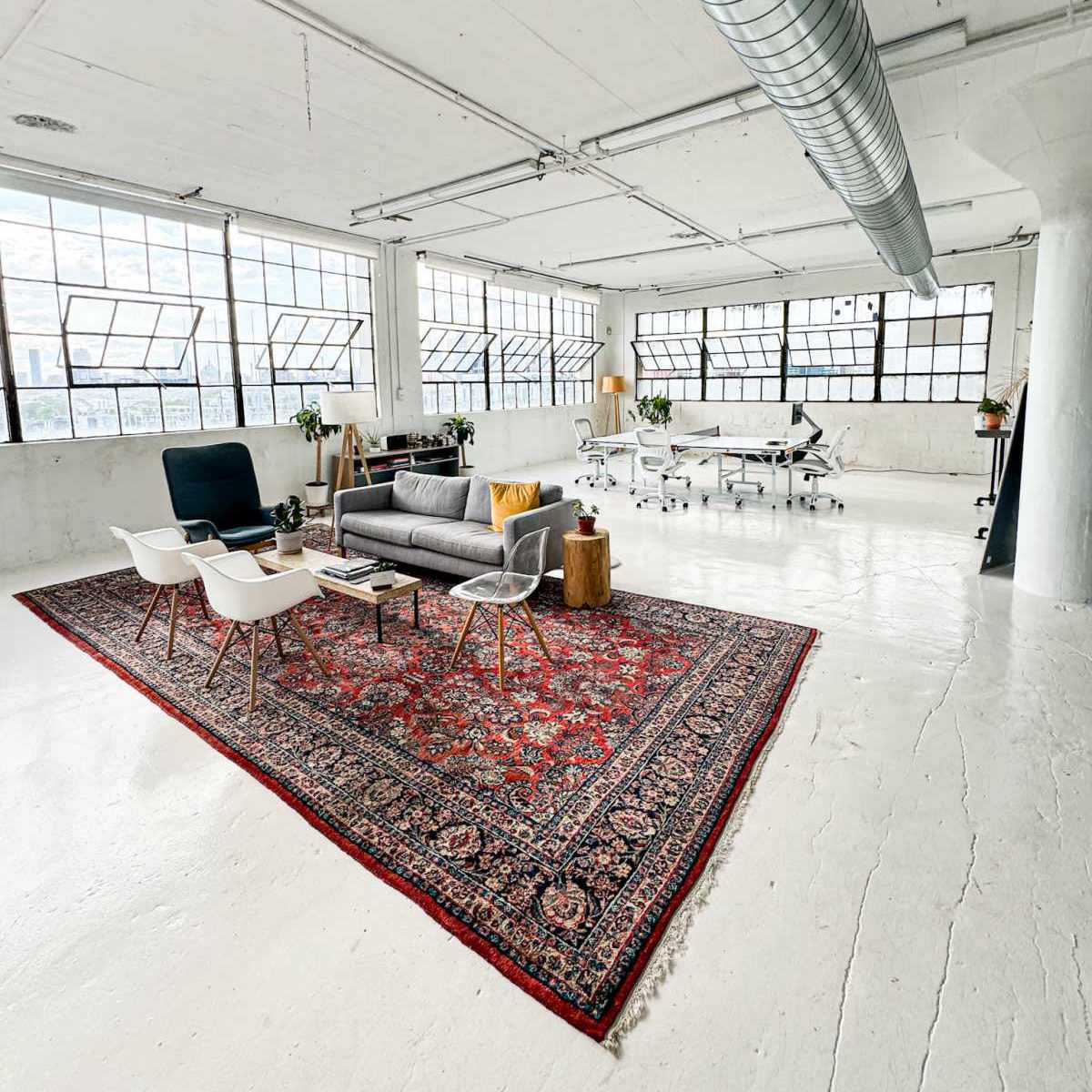 A bright, open office space features a gray sofa and white chairs on a red patterned rug, surrounded by white walls and large windows.