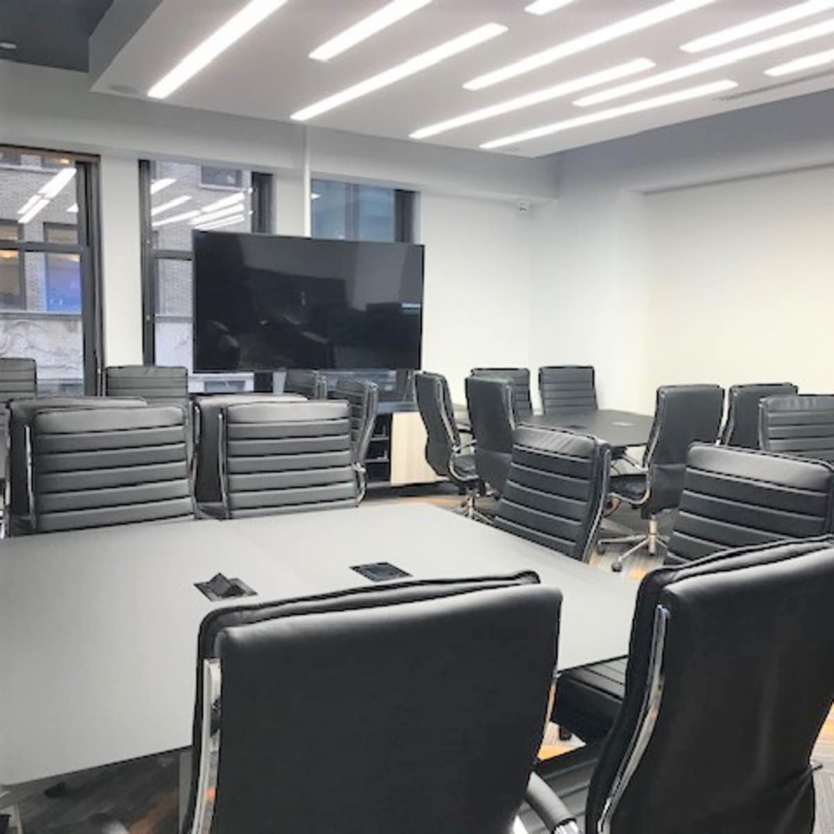 A modern conference room features a large table surrounded by several black leather chairs and a wall-mounted television.