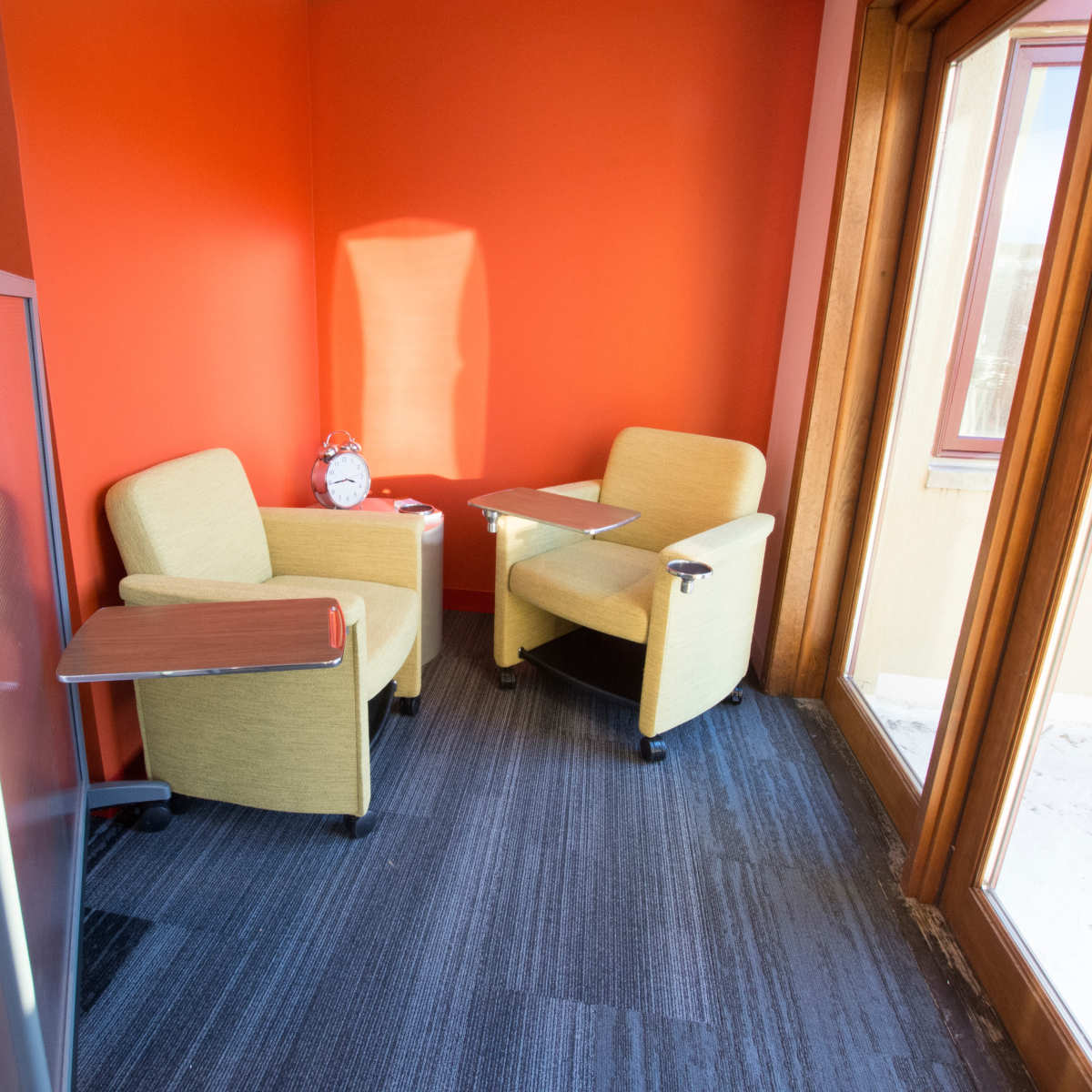 A small seating area with two yellow chairs and a wooden table against a bright orange wall, with a clock on the table and large windows letting in natural light.