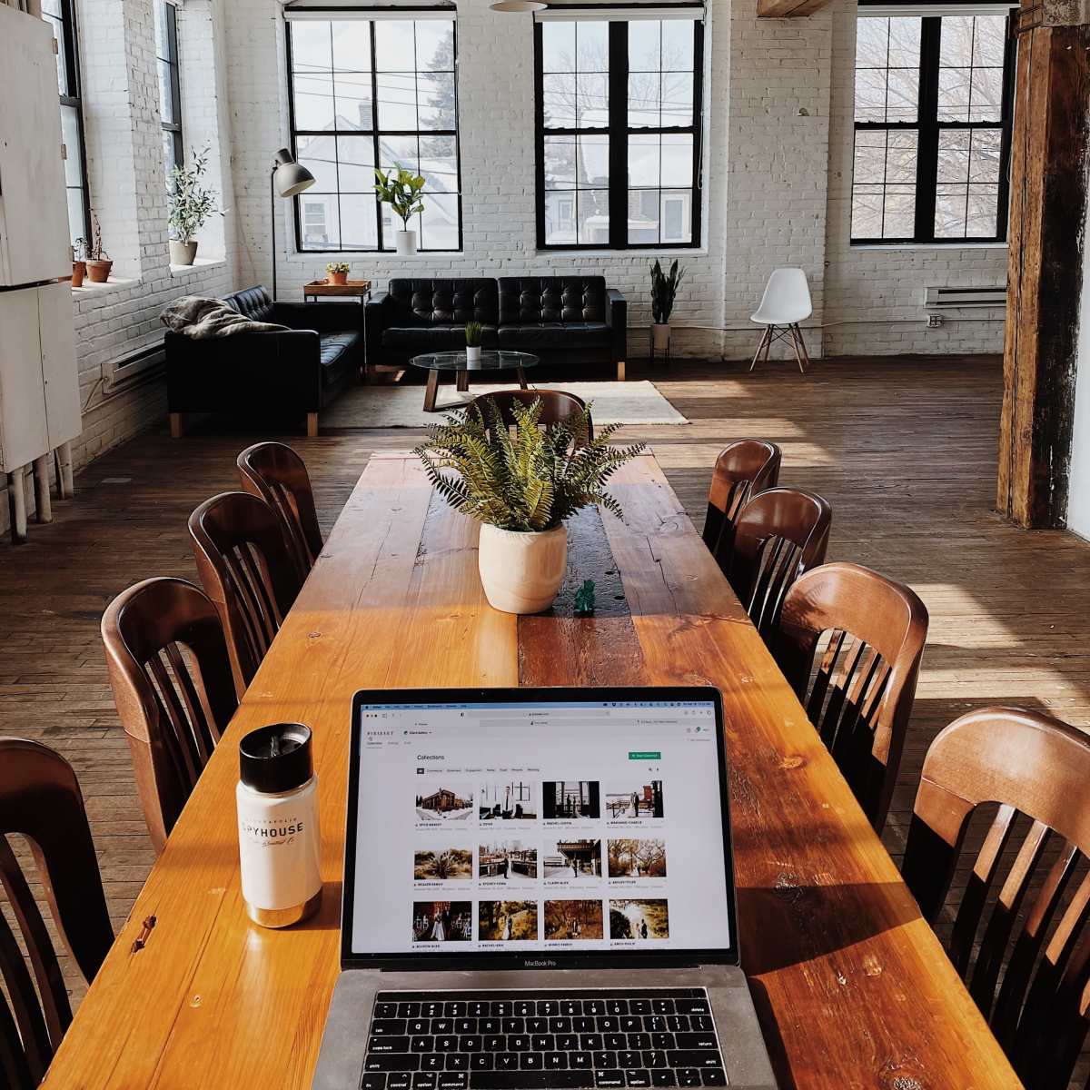 A long wooden table with a laptop open to a webpage is set in a bright, spacious room featuring large windows and modern furnishings.