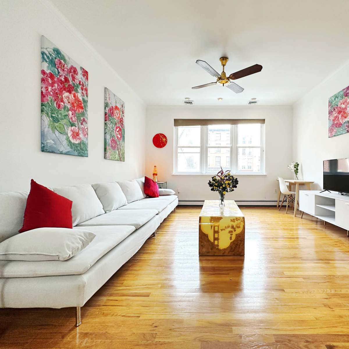 A bright living room featuring a white sectional sofa, a wooden coffee table, a TV stand with a television, and floral artwork on the walls.