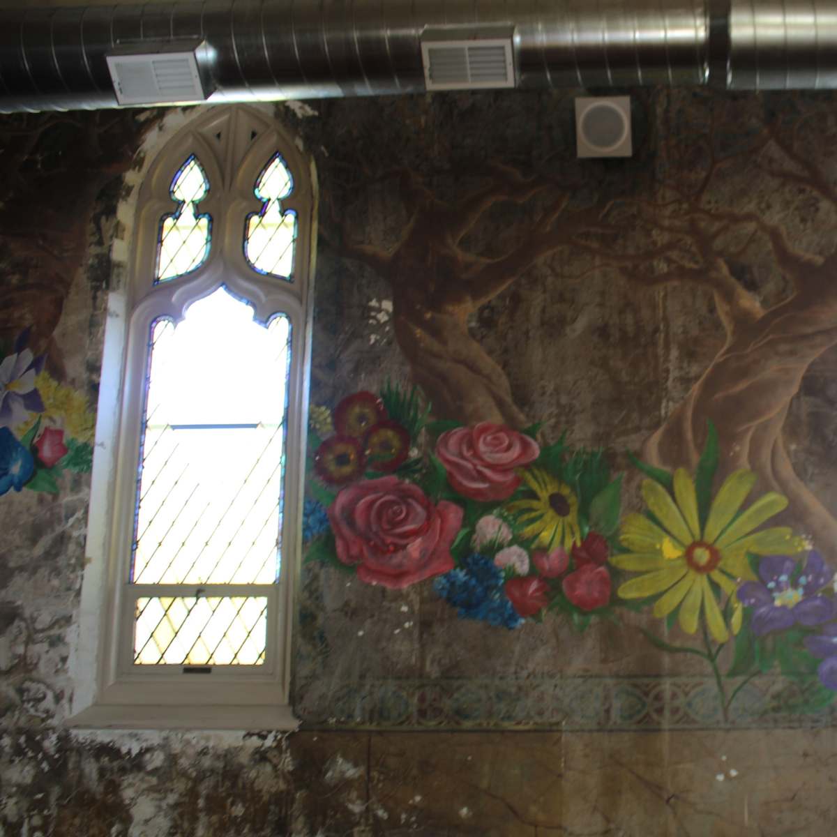 A wall with a faded mural featuring flowers and trees, flanked by arched windows with decorative glass.