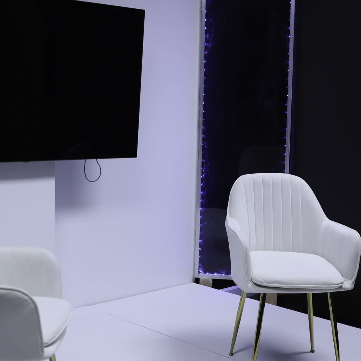 A minimalist interview setting featuring two white chairs facing a large black television screen on a light-colored backdrop.