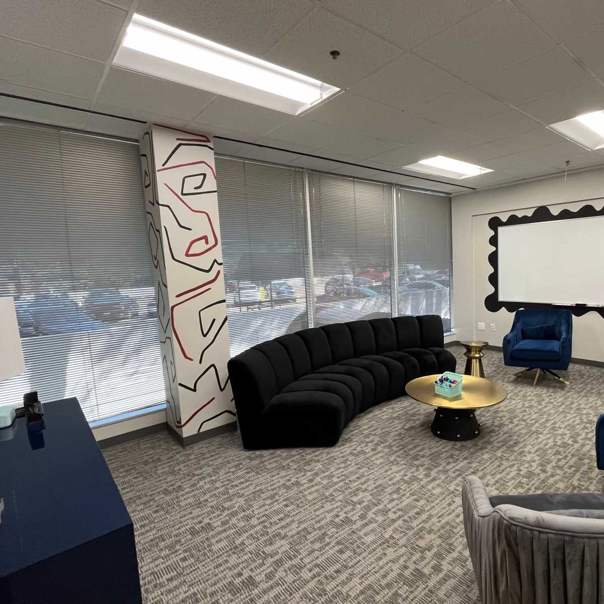 A modern office lounge features a curved black sofa, blue chairs, a round gold coffee table, and a large whiteboard against a wall.