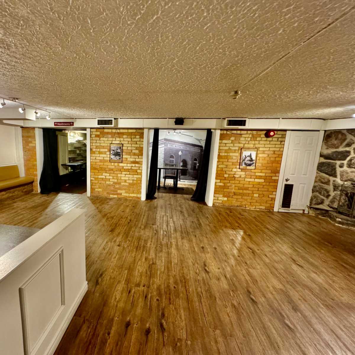 A spacious interior room with wooden flooring, brick walls, and a seating area on one side, leading to a doorway draped with curtains.