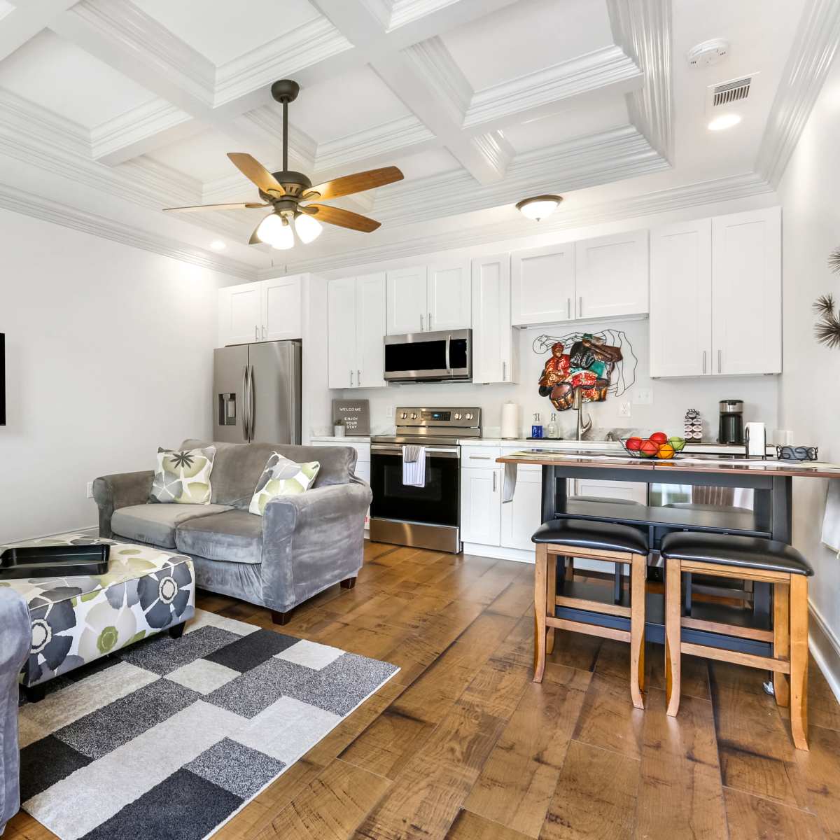 A modern living area with a gray sectional sofa, a small dining table with black stools, and a kitchen featuring white cabinets and stainless steel appliances.