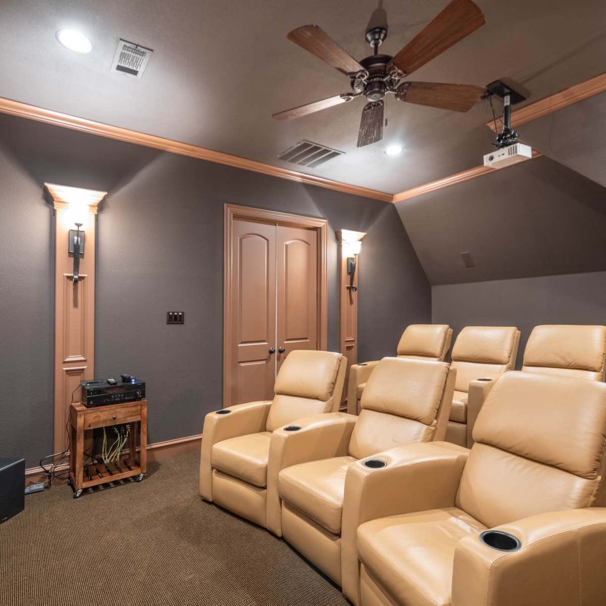 A home theater room with leather reclining seats arranged in front of a projection screen, accompanied by a ceiling fan and wall sconces illuminating the space.