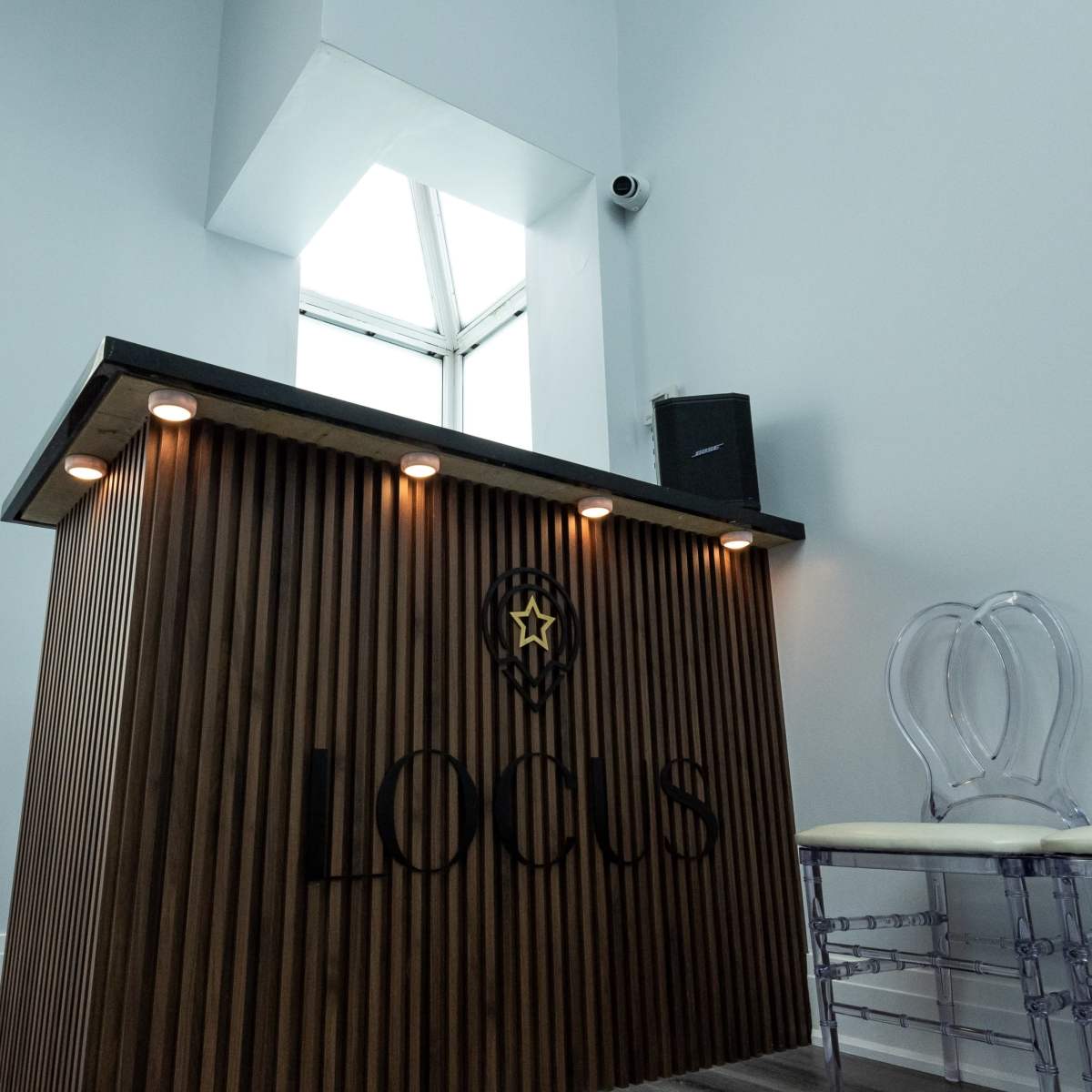 A modern reception desk with a wood panel design and the word "Locus" displayed, positioned next to clear acrylic chairs and a window above.