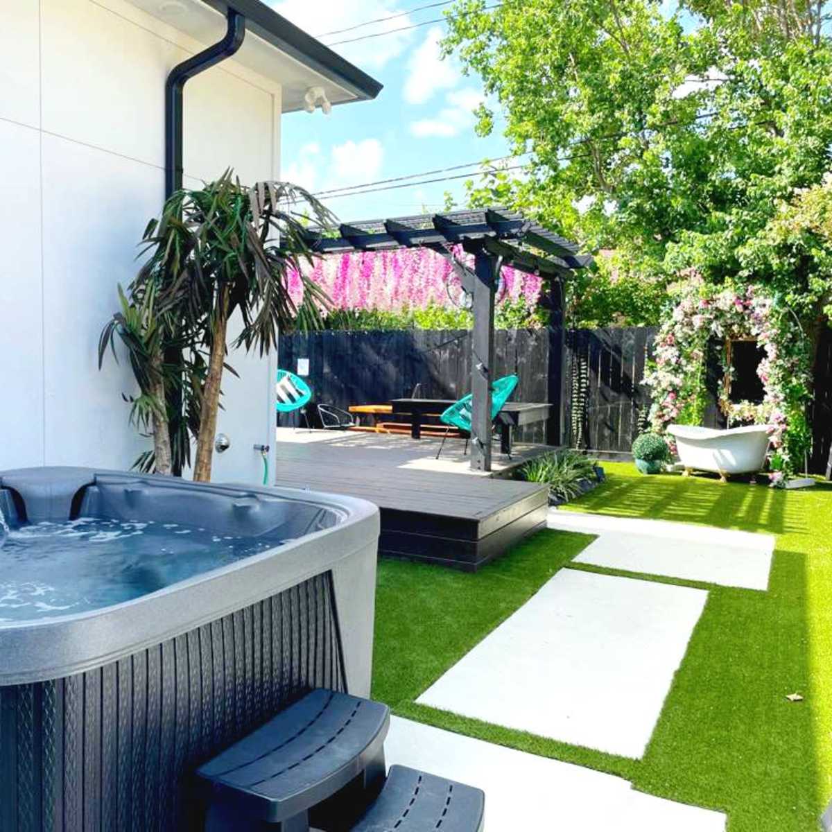 A hot tub is situated next to a deck surrounded by green grass and flowering plants in a backyard setting.