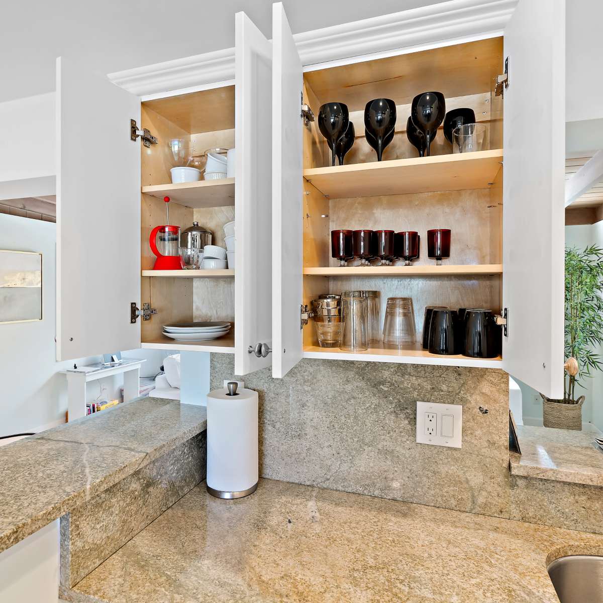 A kitchen with open cabinets displaying various glassware and dishes, along with a granite countertop and a doorway in the background.