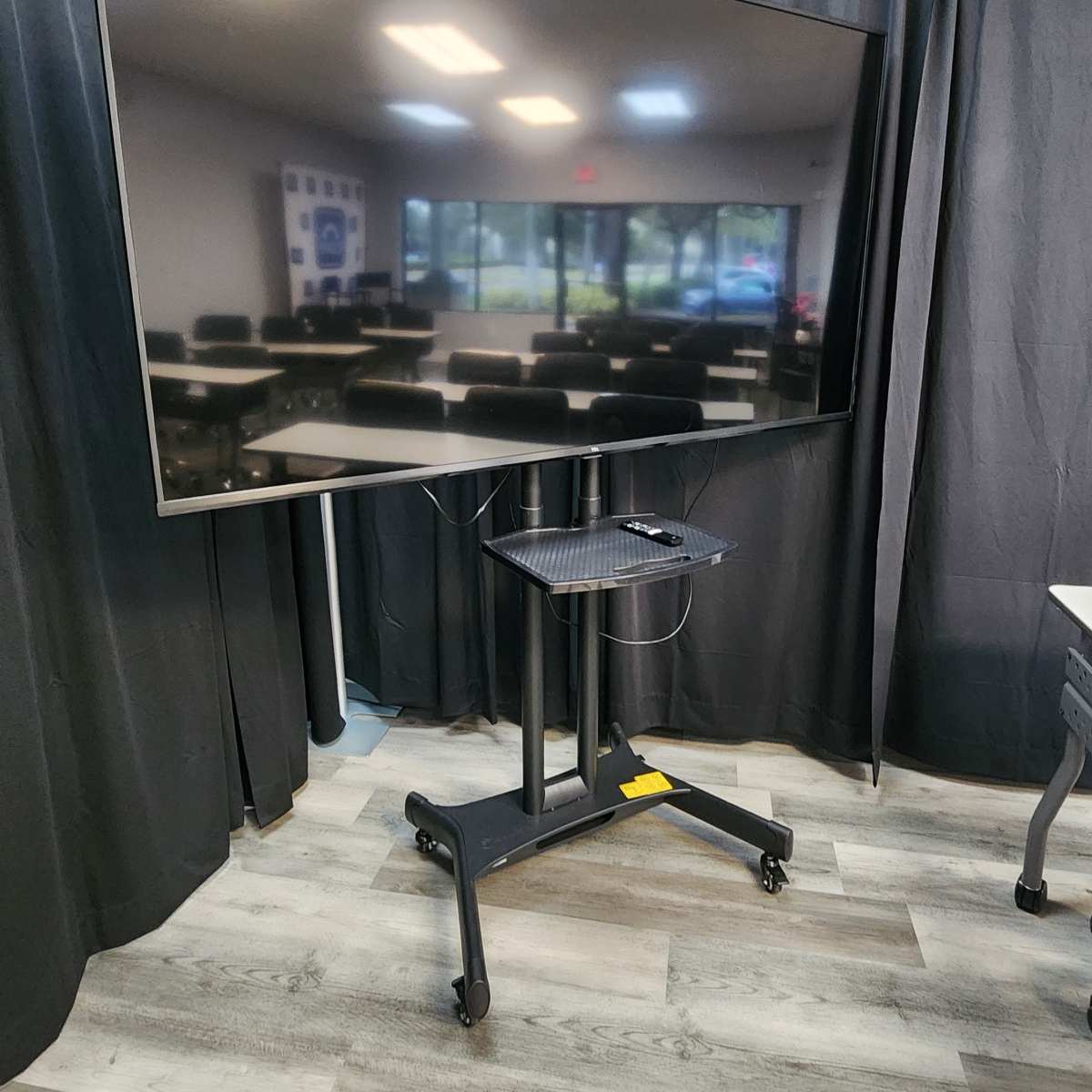 A large flat-screen monitor is mounted on a mobile stand in a room with black curtains and a view of chairs and tables in the background.
