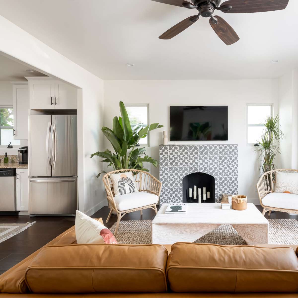 A modern living room features a brown leather couch, two rattan chairs, a coffee table, and a patterned fireplace, with a television mounted on the wall and large windows opening to greenery outside.