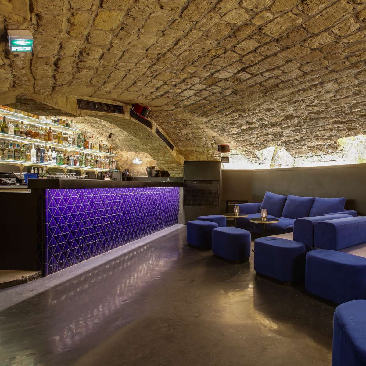 A dimly lit bar features a curved stone ceiling, a purple-lit backlit bar, and plush blue seating arrangements.