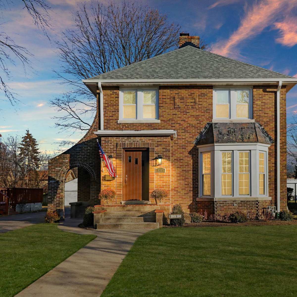 A two-story brick house with a front porch and a landscaped yard is set against a colorful sunset sky.