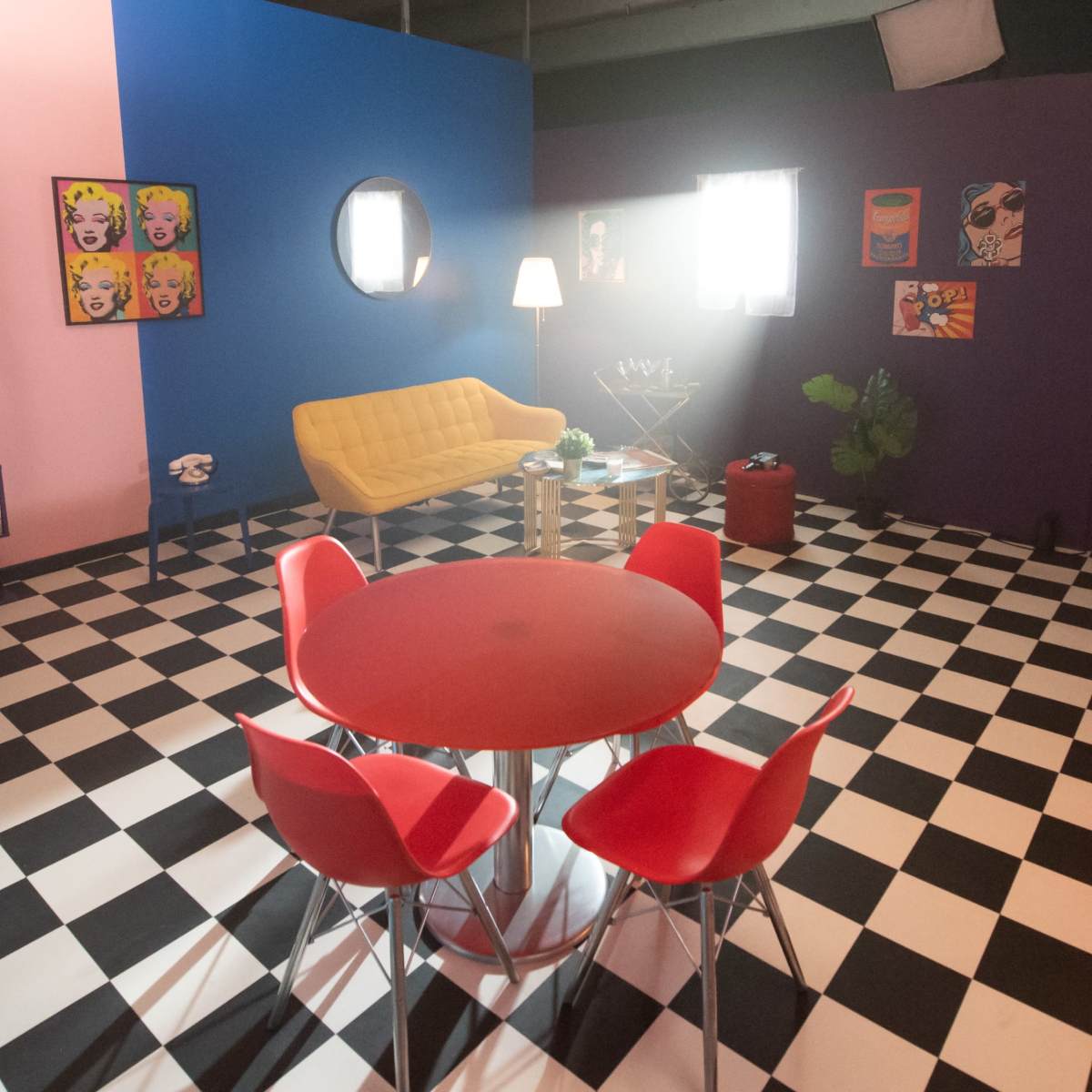 The room features a checkered black and white floor, a yellow sofa, a round red dining table surrounded by red chairs, and colorful wall decorations.