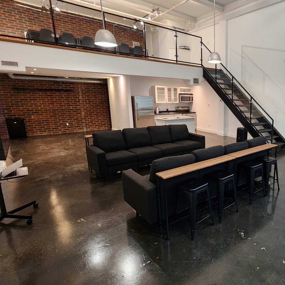 A spacious room featuring a black sectional sofa, a dining table with black chairs, a kitchen area in the background, and a staircase leading to a second level.
