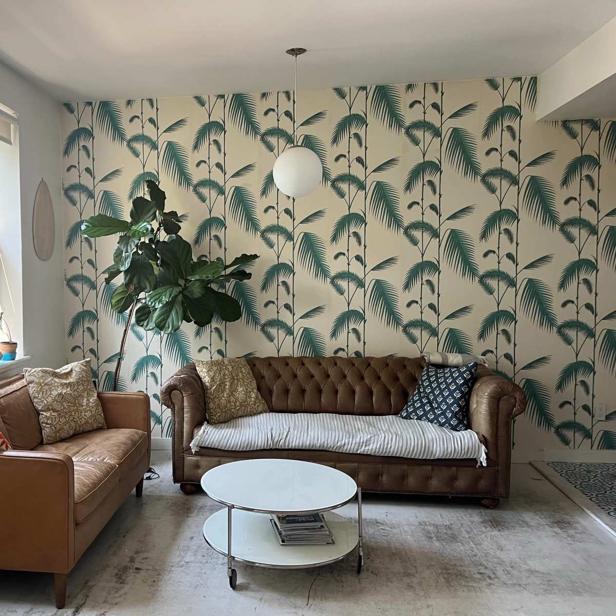 A living room with two sofas, one brown leather and the other a lighter fabric, surrounded by green plants and a patterned wallpaper featuring palm leaves.