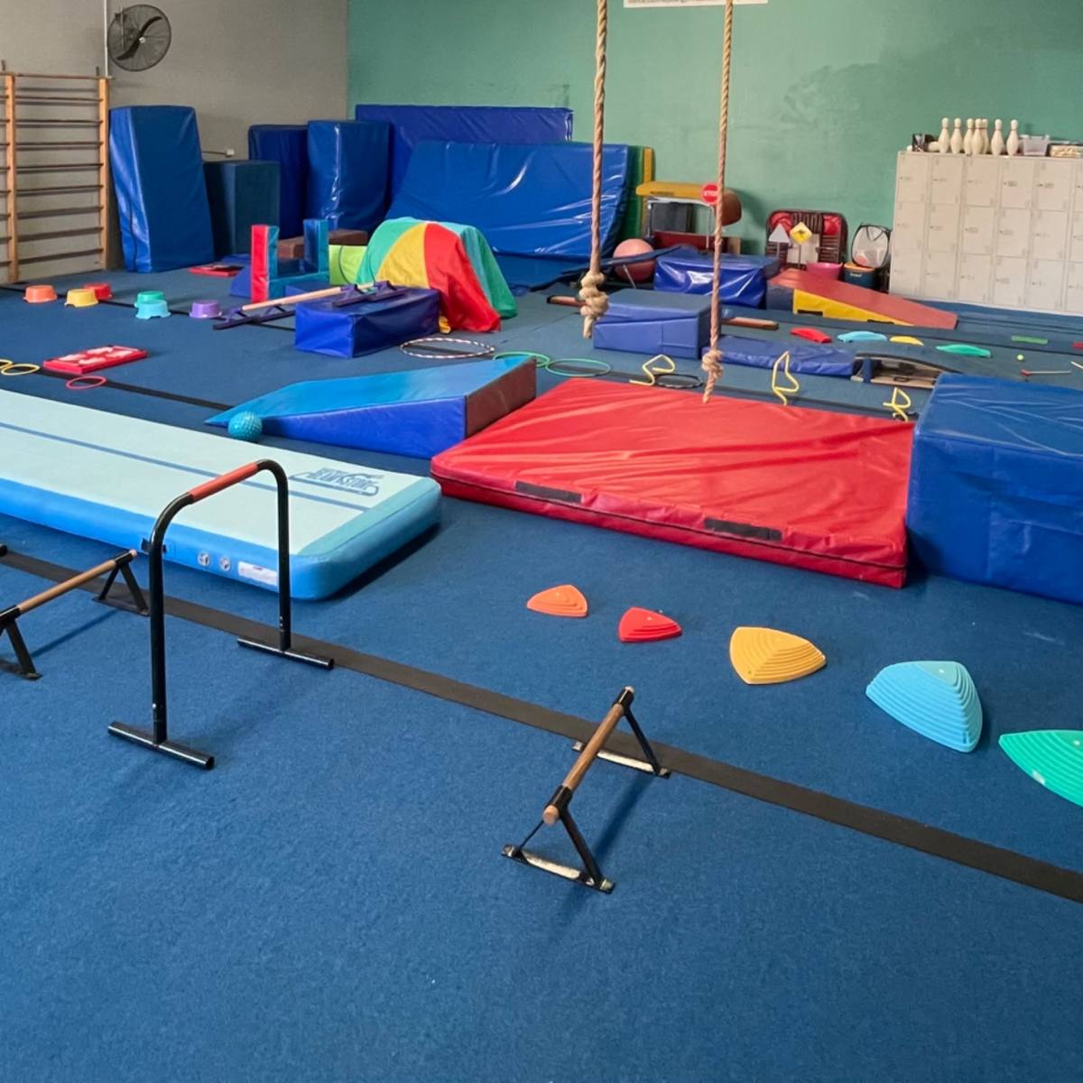 A gymnasium with various colorful gymnastic equipment, including mats, balance beams, and obstacle course elements, arranged across the blue floor.