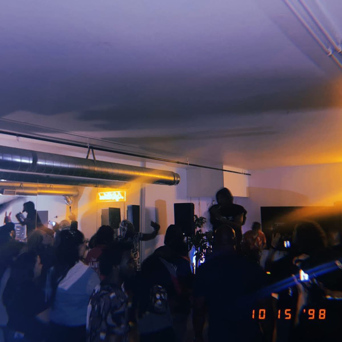 A crowd of people dances in a spacious indoor venue, with colorful lighting and a performer on stage.