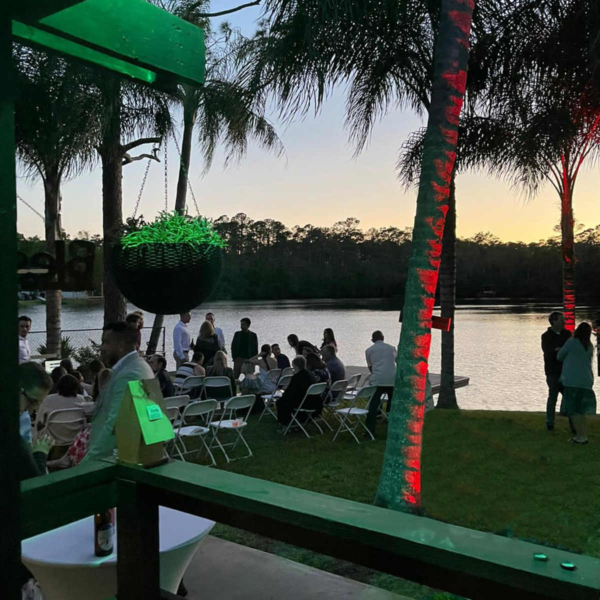 A group of people gathers along the shore of a lake at sunset, with palm trees and chairs arranged for an event.