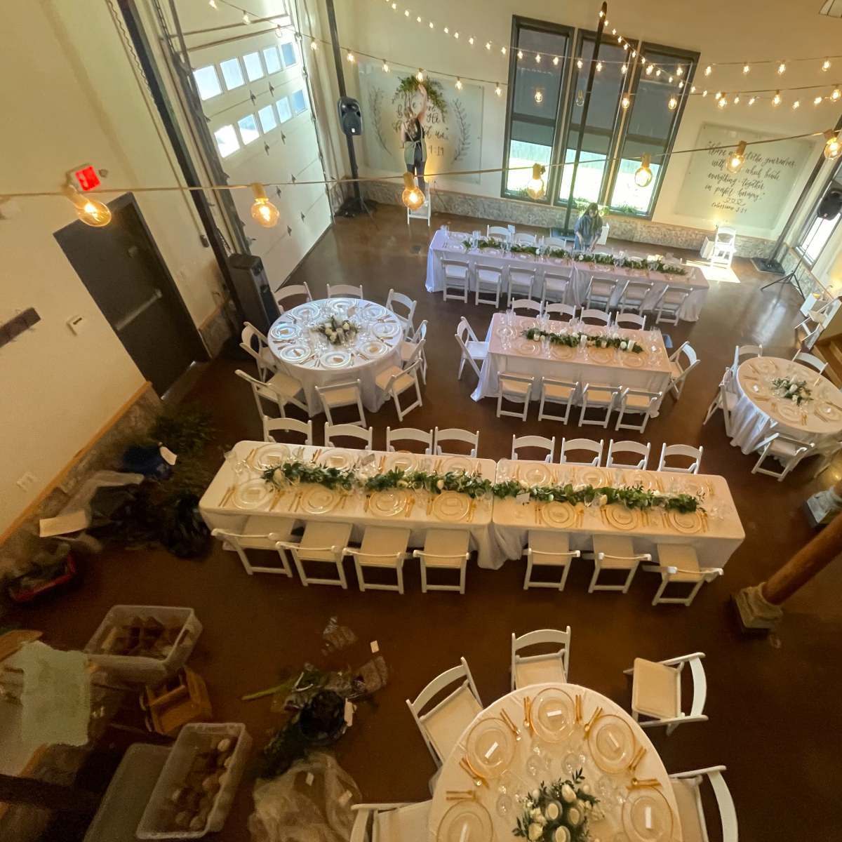 A spacious event hall arranged with multiple circular and rectangular tables decorated with white tablecloths and floral centerpieces, illuminated by string lights.