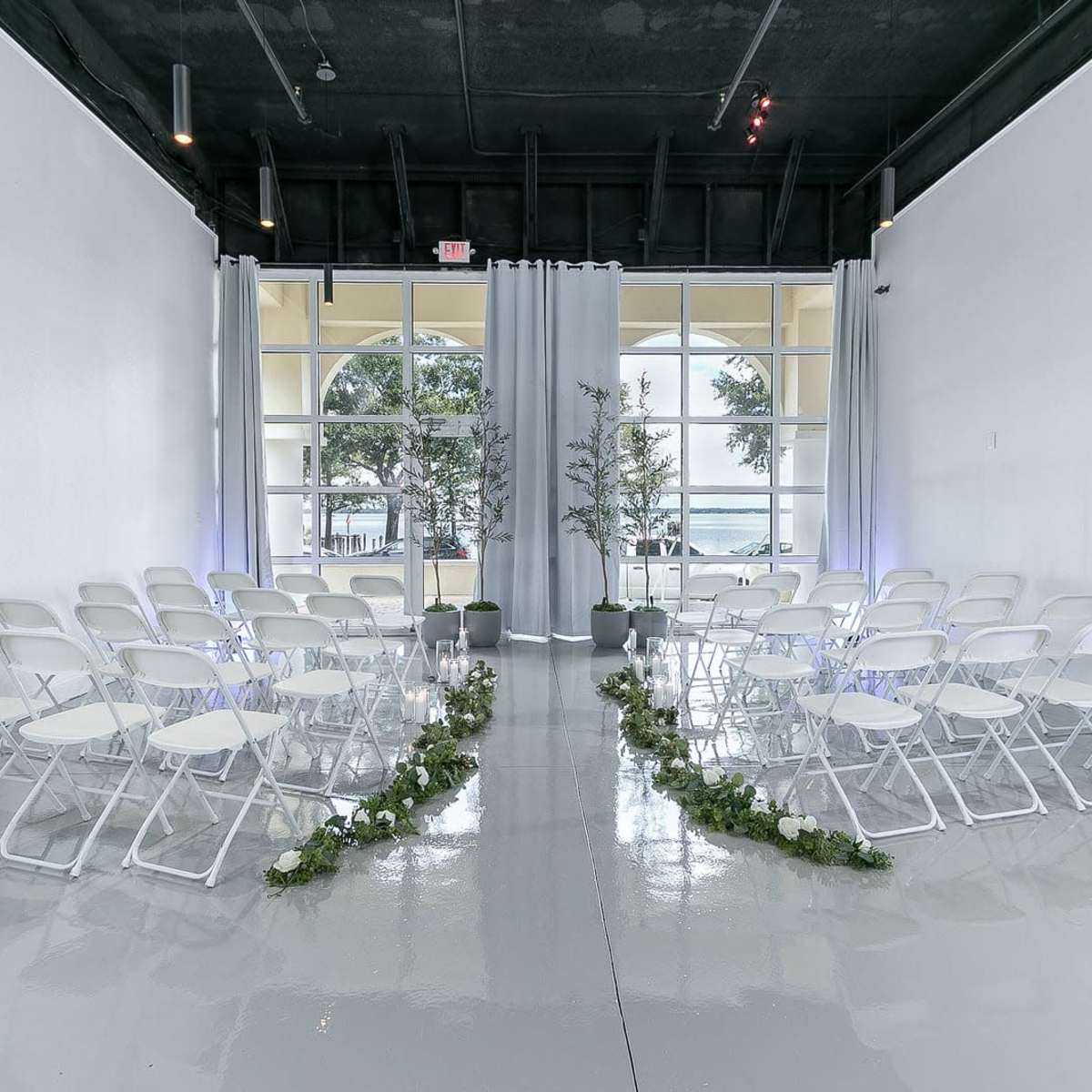 A wedding ceremony setup with white chairs arranged in two rows facing a draped area, surrounded by greenery and floral decorations.