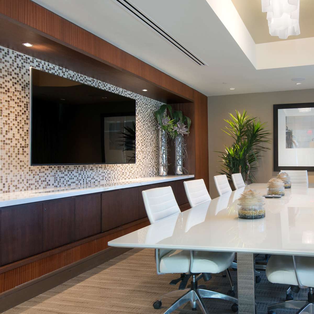 A modern conference room features a long white table surrounded by ergonomic chairs, with a decorative wall, large television, and a window providing natural light.