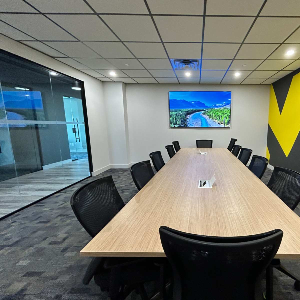 A modern conference room with a long wooden table surrounded by black chairs and a large screen displaying a landscape on the wall.