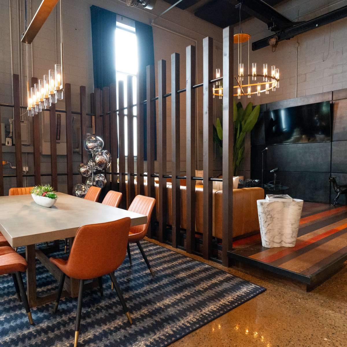 A modern dining area features a large table surrounded by orange chairs, with a partition that separates the space from a cozy living area equipped with a television and decorative elements.