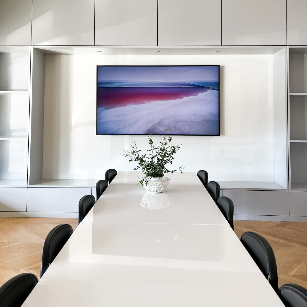 A modern conference room features a long white table with black chairs and a television mounted on a sleek gray cabinetry wall.