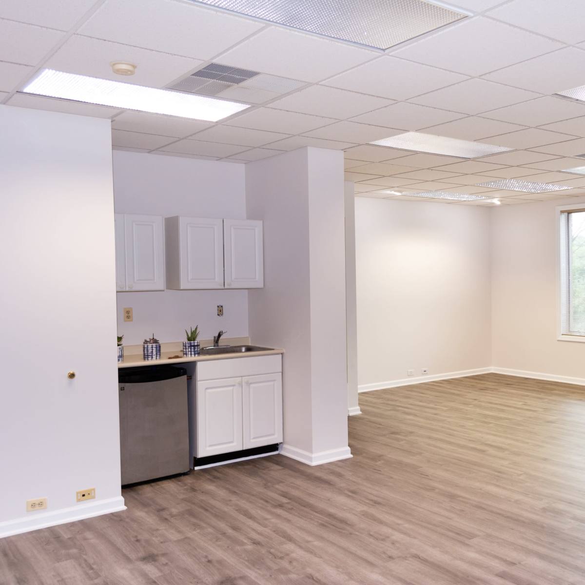 A spacious, empty room with light-colored walls, a small kitchenette in one corner, and a large window allowing natural light.