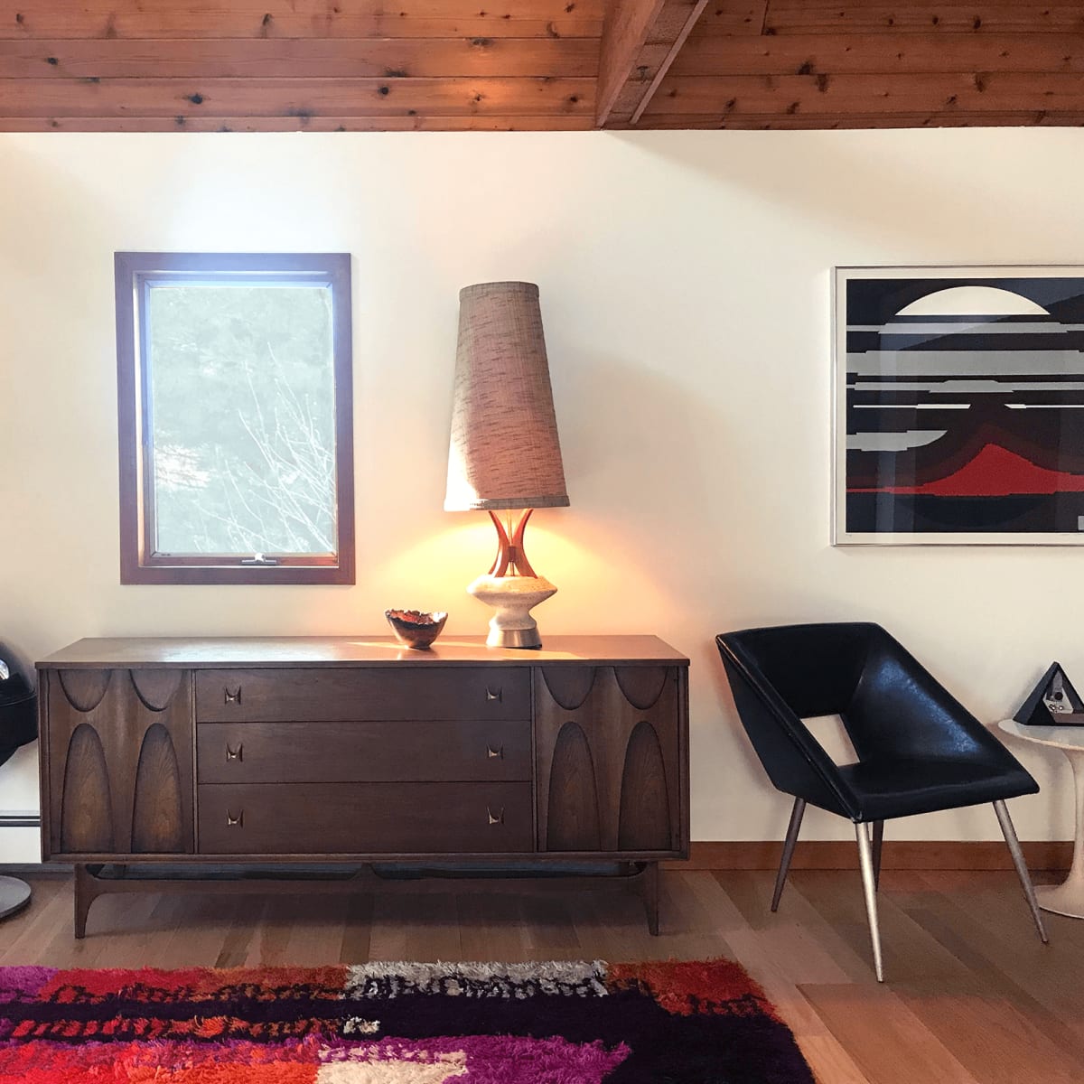 A mid-century modern interior with a wooden dresser, a tall lamp, a round black chair, and a colorful rug against a wall with a window and framed artwork.