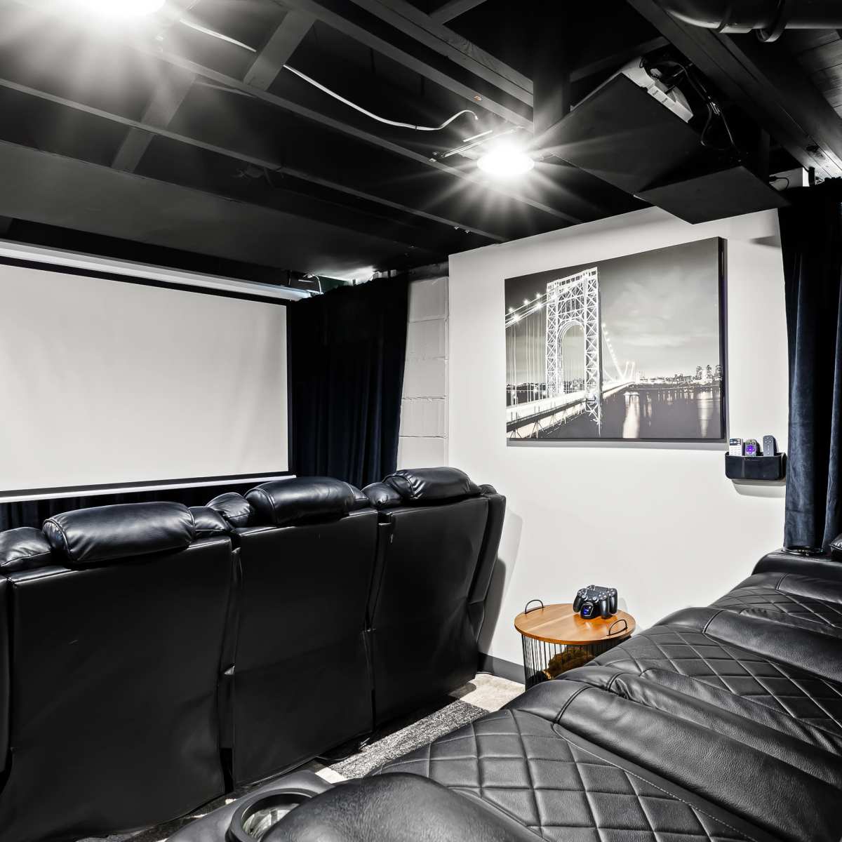 A darkened home theater room with black leather seating, a projector screen, and a wall-mounted photograph of a bridge.