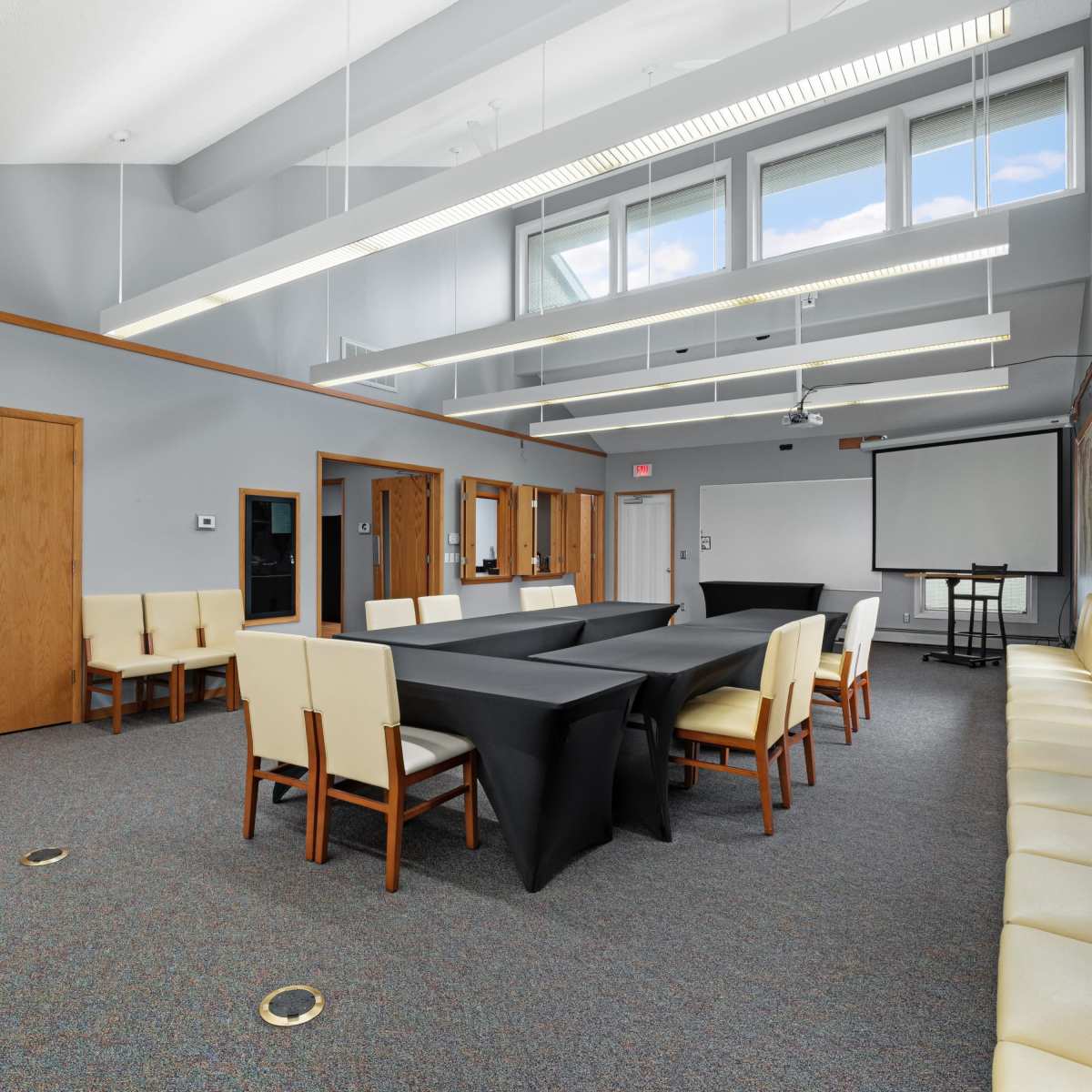 A conference room features a large black table surrounded by white chairs, with additional seating along the walls and large windows allowing natural light.