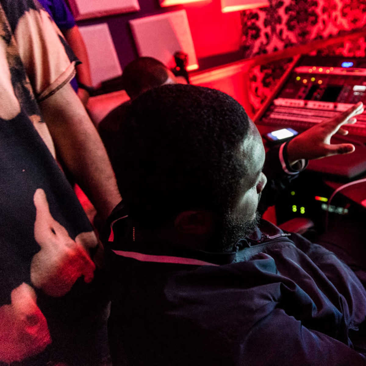 A man gestures while seated at a desk in a music recording studio surrounded by equipment and dim red lighting.