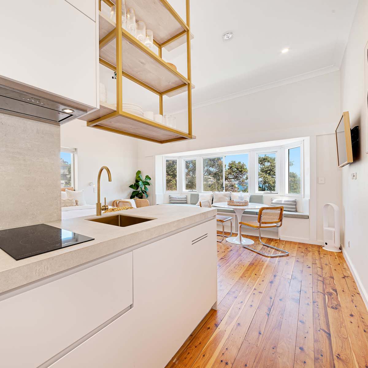 A modern kitchen features light-colored cabinetry, a sleek countertop, and a hanging display shelf, with a view of a bright dining area in the background.