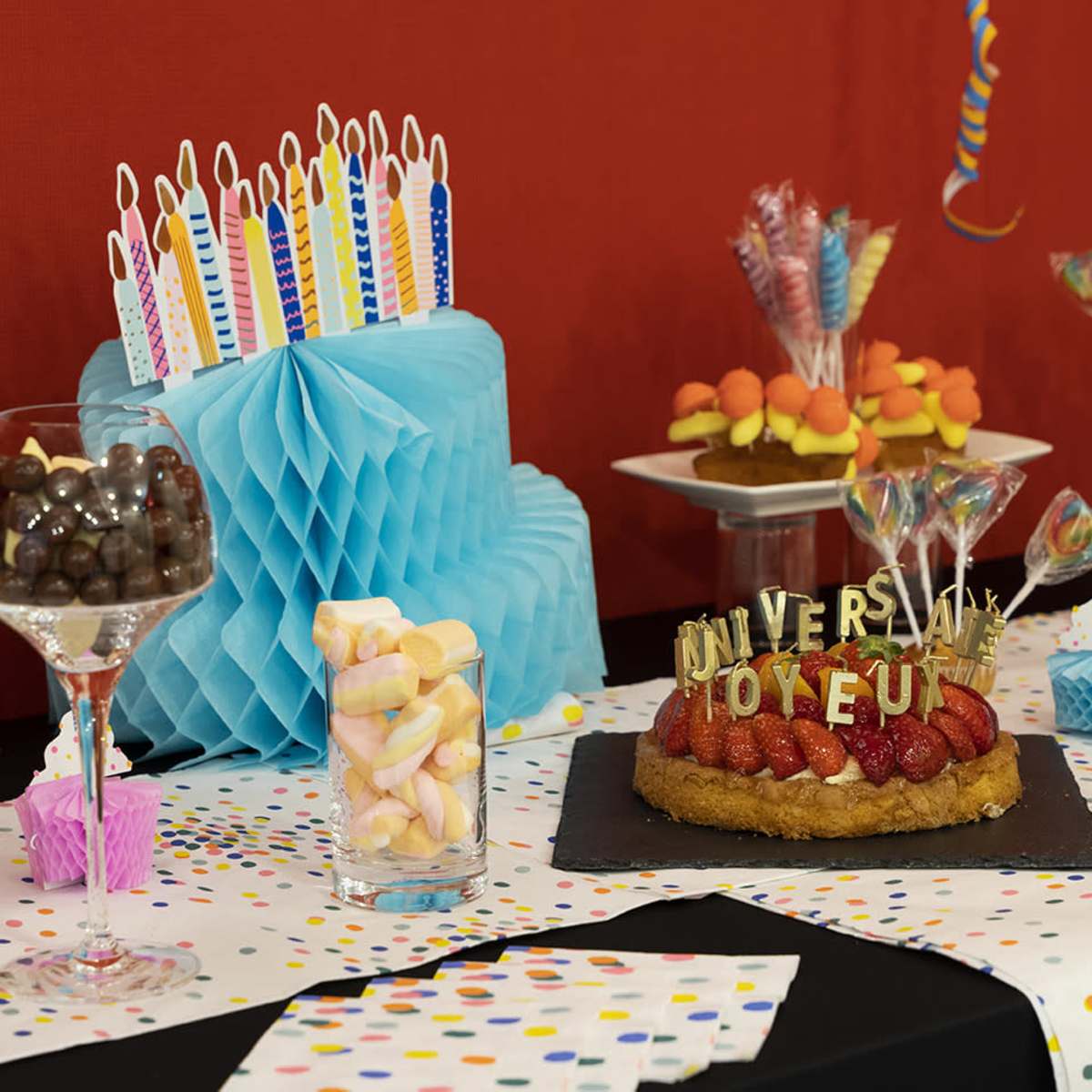 A colorful birthday party table displays a cake with decorative candles, candy-filled glasses, and various sweets arranged on a polka-dotted tablecloth.