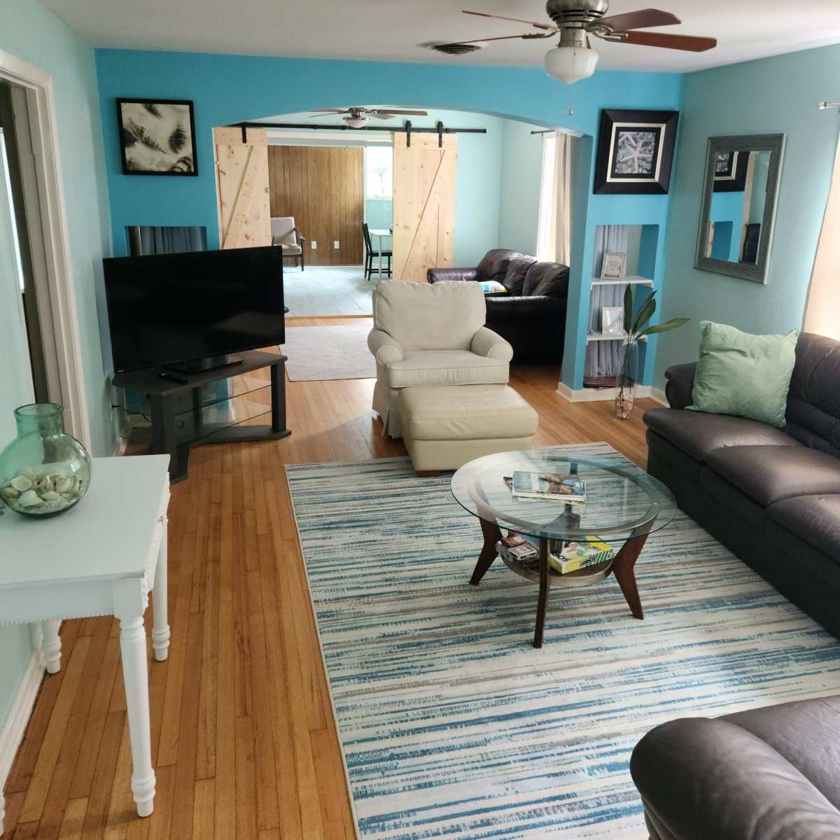 A living room features a light blue wall, a television, two couches, a coffee table, and a view of a room beyond with sliding barn doors.